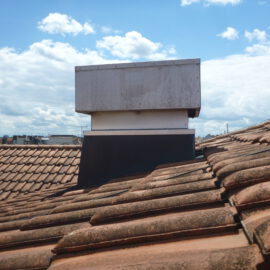 Das Bild zeigt einen rechteckigen Luftauslass auf einem geneigten Dach mit roten Dachziegeln. Der Auslass besteht aus einem unteren schwarzen Sockel, einem weißen Mittelteil und einem oberen silberfarbenen Metallgehäuse, das leicht verwittert wirkt. Im Hintergrund sind weitere Dächer und eine Stadtlandschaft unter einem blauen Himmel mit weißen Wolken zu sehen.