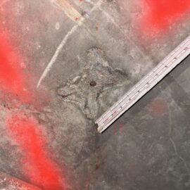 Das Bild zeigt eine Betonoberfläche, die teilweise mit roter Markierungsfarbe besprüht ist. In der Mitte befindet sich ein metallischer Abstandshalter, der sternförmig im Beton eingebettet ist und eine kleine runde Öffnung in der Mitte aufweist. Rechts im Bild liegt ein Maßstab, der diagonal über die Fläche verläuft und zur Größenbestimmung dient. Die Oberfläche des Betons ist rau und weist Schleifspuren sowie leichte Verschmutzungen auf.