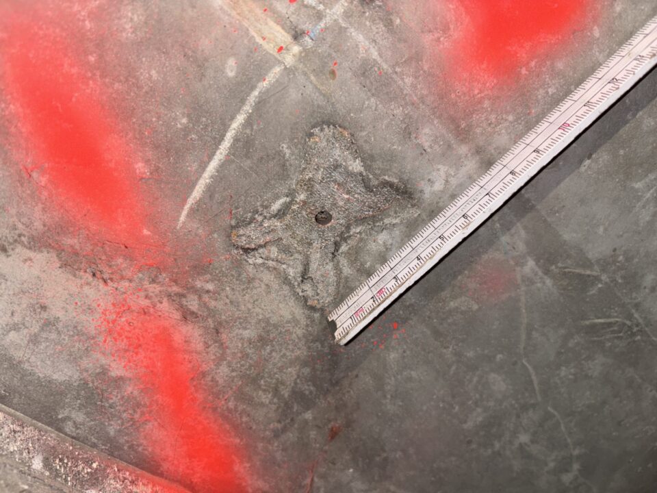 Das Bild zeigt eine Betonoberfläche, die teilweise mit roter Markierungsfarbe besprüht ist. In der Mitte befindet sich ein metallischer Abstandshalter, der sternförmig im Beton eingebettet ist und eine kleine runde Öffnung in der Mitte aufweist. Rechts im Bild liegt ein Maßstab, der diagonal über die Fläche verläuft und zur Größenbestimmung dient. Die Oberfläche des Betons ist rau und weist Schleifspuren sowie leichte Verschmutzungen auf.