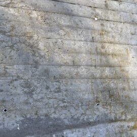Das Bild zeigt eine grobe Betonwand mit horizontalen Schalungsabdrücken, die auf eine gegossene Bauweise hinweisen. Die Oberfläche ist uneben und weist Verfärbungen sowie Spuren von Witterungseinflüssen auf. Mehrere kleine runde Öffnungen sind in regelmäßigen Abständen in der Wand eingelassen, vermutlich für Befestigungen oder zur Kontrolle der Mauerstärke. Die Farbgebung ist überwiegend grau mit leichten gelblichen Ablagerungen.