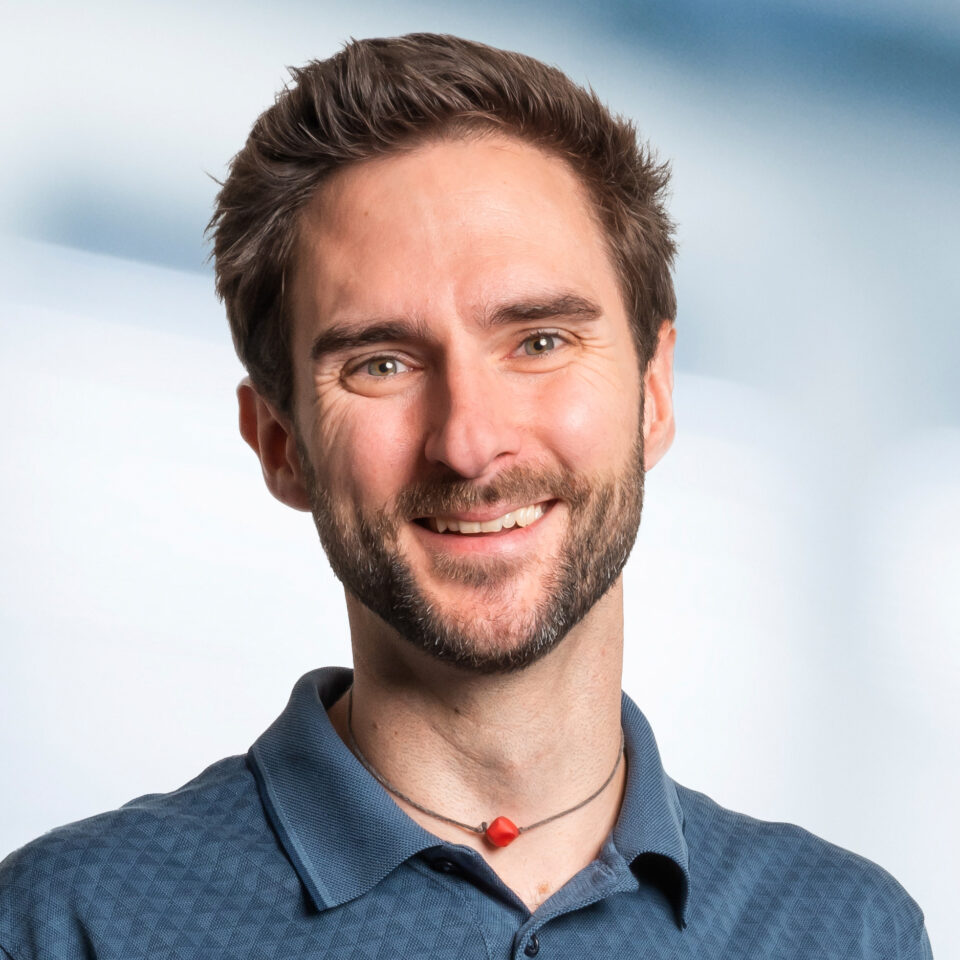 Das Bild zeigt ein freundlich lächelndes Porträt einer Person mit kurzem braunen Haar und gepflegtem Bart. Die Person trägt ein blaues Polohemd sowie eine Kette mit einem kleinen roten Anhänger. Der Hintergrund ist hell und neutral gehalten, wodurch das Gesicht klar im Fokus steht.