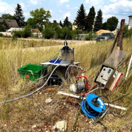 Das Bild zeigt einen messtechnischen Aufbau auf einer offenen, grasbewachsenen Fläche. Zu sehen sind verschiedene Geräte und Materialien, darunter ein grüner Kunststoffbehälter, ein Metallgestell mit Schläuchen und Kabeln, eine blaue Kabeltrommel sowie ein Steuerkasten. Im Hintergrund befinden sich Häuser, Bäume und ein teilweise bewölkter Himmel, was auf eine ländliche Umgebung hinweist.