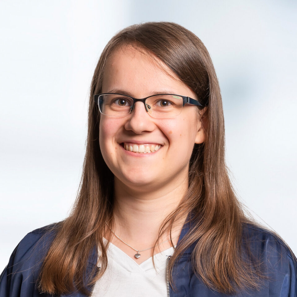 Das Bild zeigt ein professionelles Porträt einer lächelnden Frau mit langen, braunen Haaren, die zur Seite gescheitelt sind. Sie trägt eine dunkle Brille mit rechteckigem Rahmen, ein weißes Oberteil und darüber eine dunkelblaue Jacke. Um den Hals trägt sie eine feine Kette mit einem herzförmigen Anhänger.