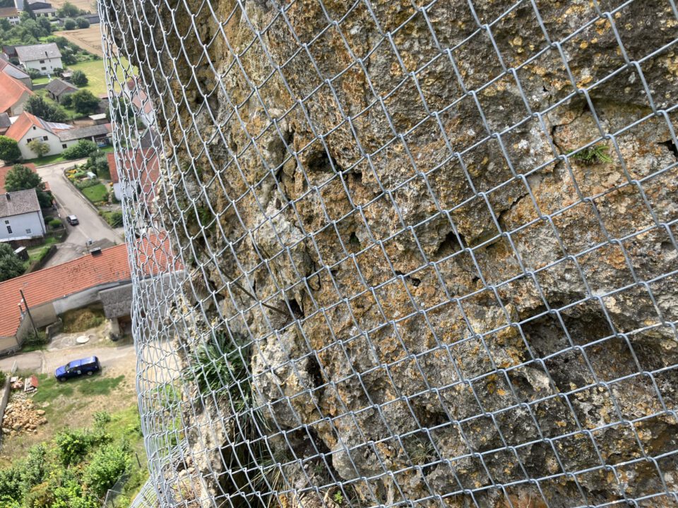 Das Bild zeigt eine steile Felswand, die mit einem engmaschigen Drahtnetz gesichert ist. Das Netz liegt dicht an der unregelmäßigen, bräunlich-grauen Felsoberfläche an, auf der vereinzelt kleine Pflanzen wachsen. Im Hintergrund sind Dächer von Häusern und ein blauer Transporter auf einer Straße zu erkennen, was auf eine ländliche Umgebung hinweist.