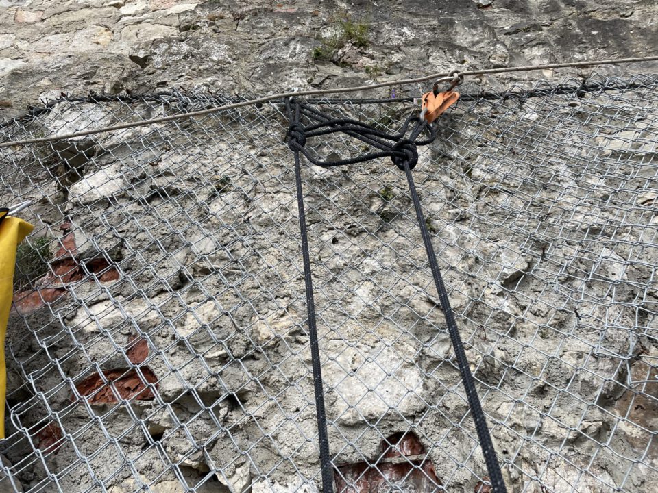 Das Bild zeigt eine Felswand, die mit einem stabilen Drahtnetz gesichert ist. Am oberen Rand ist das Netz mit einem Stahlseil befestigt, an dem ein Knoten und eine Metallverbindung zu sehen sind. Im Vordergrund hängen zwei schwarze Seile am Netz, und links ist ein Teil einer gelben Tasche erkennbar.