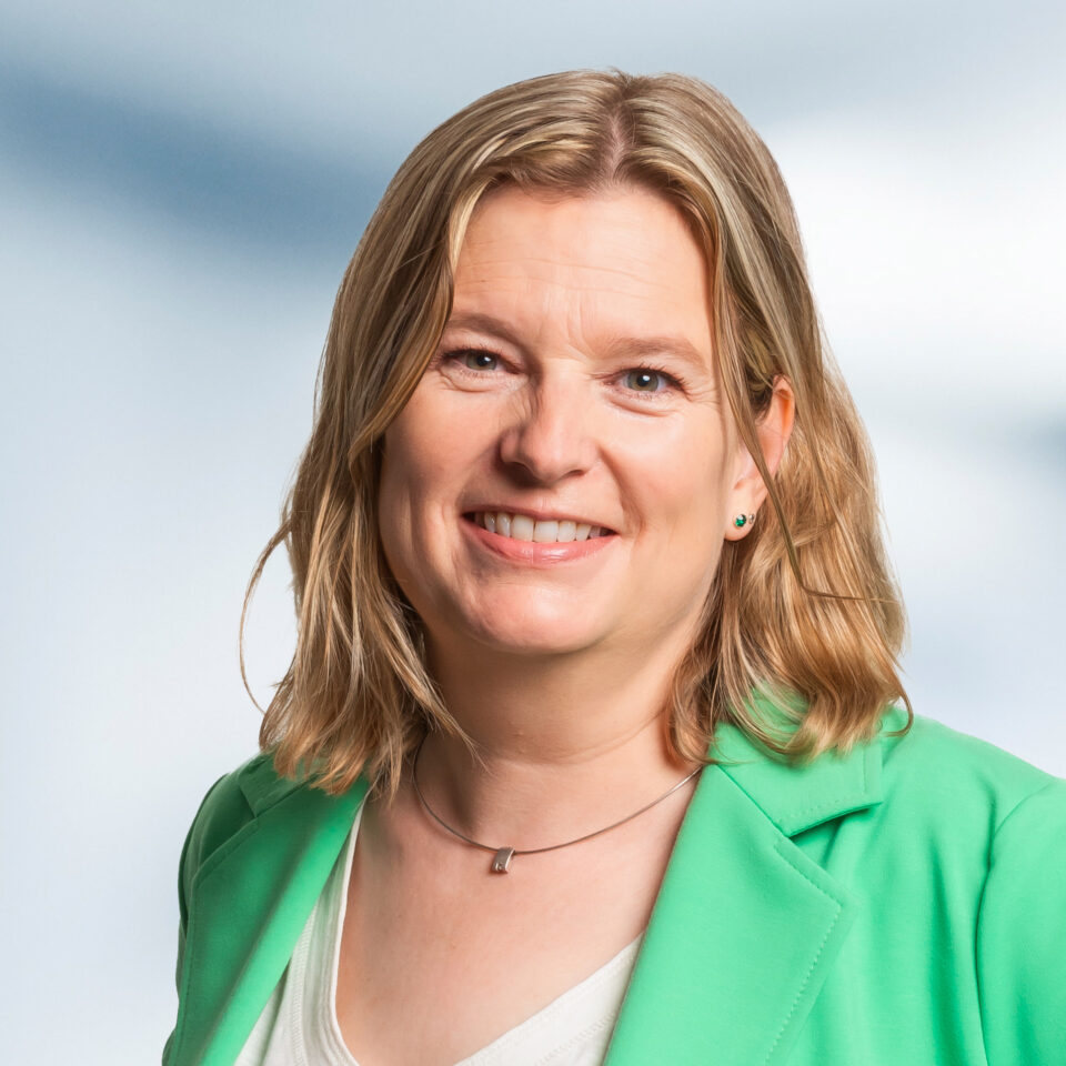 Das Bild zeigt ein professionelles Porträt einer Frau mit schulterlangen, leicht gewellten blonden Haaren. Sie lächelt freundlich in die Kamera. Sie trägt ein helles Oberteil, darüber einen grünen Blazer sowie eine feine Kette mit kleinem Anhänger. Als Schmuck sind zudem kleine, dezente Ohrstecker zu sehen. Der Hintergrund ist weich und ruhig in einem Verlauf aus hellen Blau- und Weißtönen gehalten, was dem Porträt eine helle, klare und professionelle Wirkung verleiht.