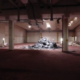 Das Bild zeigt eine große, leere Halle mit einem rötlichen Boden und hellen Wänden, die von mehreren Säulen unterteilt wird. In der Mitte liegt ein großer Haufen dunkler, zerbrochener Materialien, während im Hintergrund weitere kleinere Schutthaufen zu sehen sind, die durch Absperrbänder abgegrenzt sind. Die Beleuchtung ist gedämpft, einige Deckenlampen werfen Licht auf die zentralen Bereiche, und an der Decke verlaufen sichtbare Lüftungsrohre.