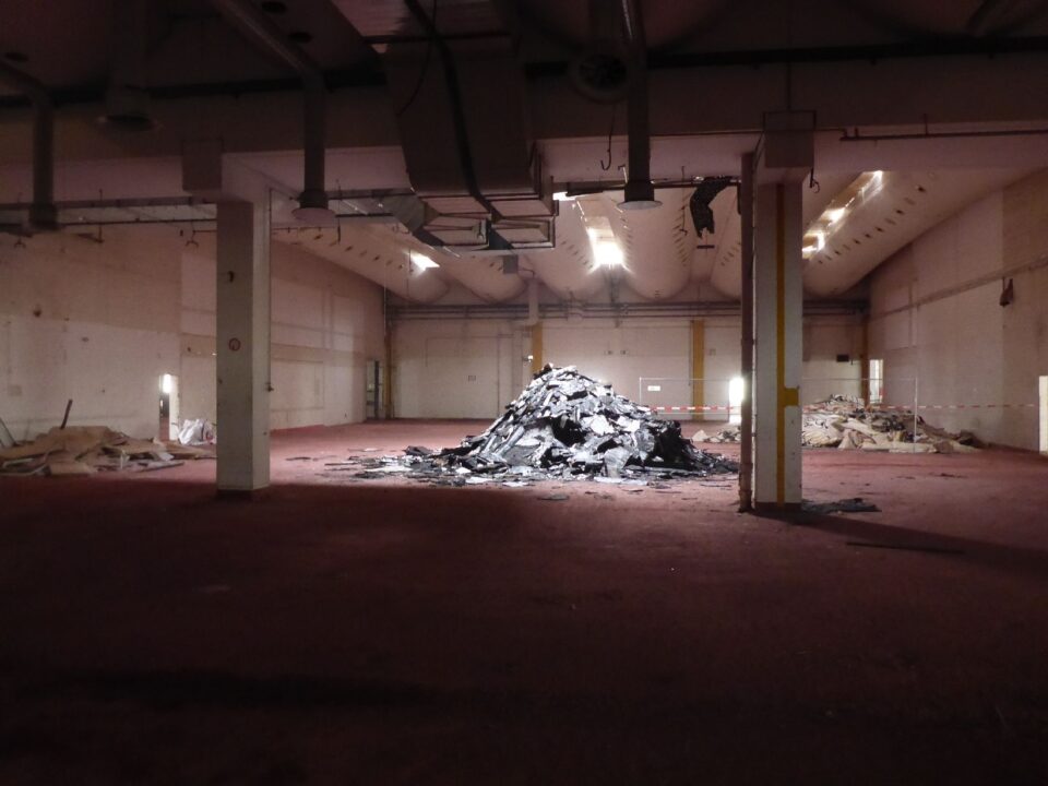 Das Bild zeigt eine große, leere Halle mit einem rötlichen Boden und hellen Wänden, die von mehreren Säulen unterteilt wird. In der Mitte liegt ein großer Haufen dunkler, zerbrochener Materialien, während im Hintergrund weitere kleinere Schutthaufen zu sehen sind, die durch Absperrbänder abgegrenzt sind. Die Beleuchtung ist gedämpft, einige Deckenlampen werfen Licht auf die zentralen Bereiche, und an der Decke verlaufen sichtbare Lüftungsrohre.