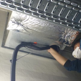 Das Bild zeigt eine Person, die an einer Lüftungsanlage arbeitet. Die Lüftungsöffnung ist mit silberner Isolierfolie verkleidet, und die Person hält einen schwarzen Schlauch in die Öffnung, vermutlich für eine Inspektion oder Reinigung. Über der Öffnung verläuft eine Metallkabeltrasse, und die Umgebung besteht aus einer hellen Wand und einer technischen Decke.