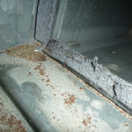 Das Bild zeigt eine Nahaufnahme einer beschädigten Dichtung in einem metallischen Lüftungskanal. Die Dichtung ist stark bröckelig und weist Risse sowie abgeplatzte Stellen auf, wodurch das Material ungleichmäßig wirkt. Auf den angrenzenden Metallflächen sind Rostpartikel und Staub verteilt, was auf Verschleiß und mangelnde Abdichtung hinweist.