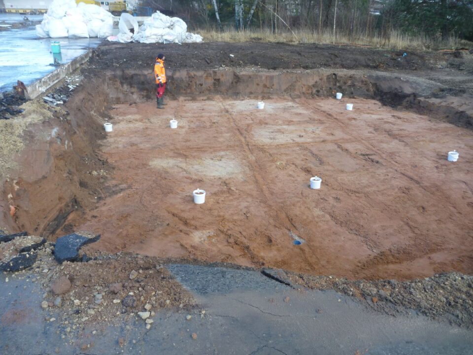 Das Bild zeigt eine rechteckige Baugrube mit freigelegtem Erdreich, auf dem mehrere weiße Eimer gleichmäßig verteilt aus dem Boden stehen. Am Rand der Grube steht eine Person in orangefarbener Sicherheitskleidung, die offenbar die Arbeiten überwacht. Im Hintergrund sind aufgeschüttete Erde, einige weiße Säcke und eine asphaltierte Fläche zu erkennen, während die Umgebung von Gras und Bäumen begrenzt wird.