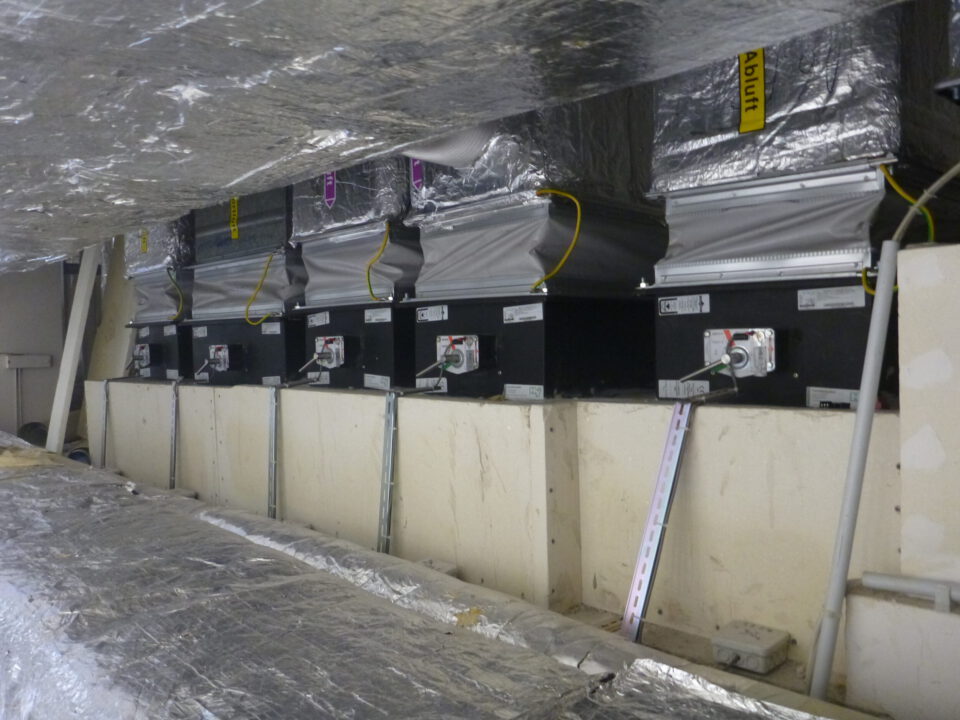 Das Bild zeigt eine technische Installation in einem Innenraum, vermutlich eine Lüftungs- oder Klimaanlage. Mehrere rechteckige Geräte sind in einer Reihe montiert, darüber verlaufen große Kanäle, die mit silberner Isolierfolie ummantelt sind. Gelbe Kabel und flexible Anschlüsse verbinden die Geräte mit den Kanälen, und an den Geräten sind Stellhebel und Beschriftungen sichtbar.