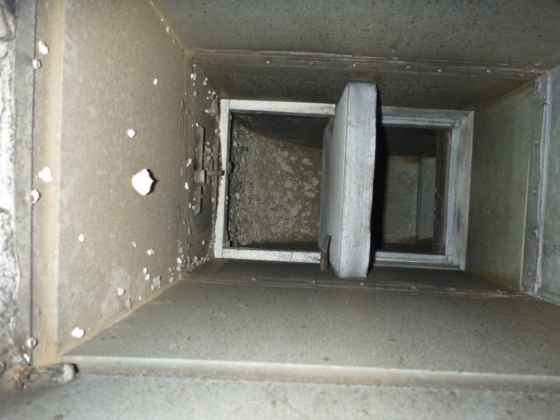 Das Bild zeigt das Innere eines rechteckigen Lüftungskanals aus Metall, der stark verschmutzt ist und Ablagerungen an den Wänden aufweist. In der Mitte befindet sich eine Klappe oder ein Schieber, der teilweise geöffnet ist und an einem Rahmen befestigt ist. Die Oberflächen sind matt und zeigen deutliche Spuren von Staub und Korrosion, was auf eine lange Nutzungsdauer oder fehlende Reinigung hinweist.