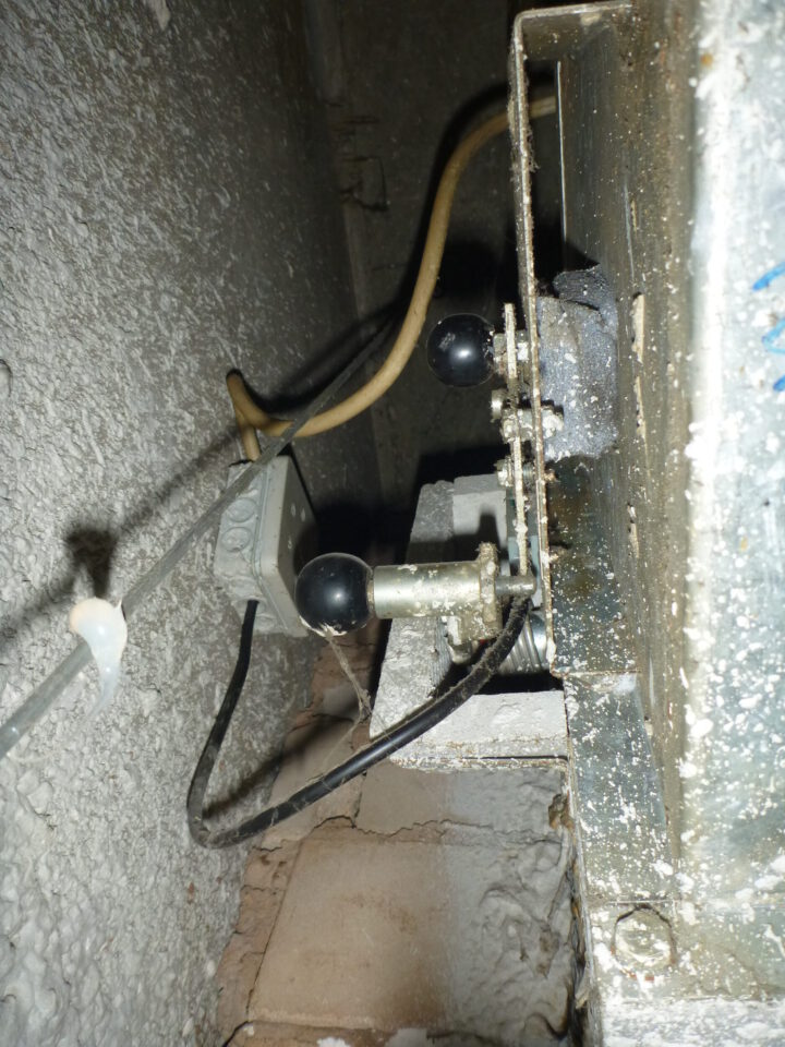 Das Bild zeigt eine Nahaufnahme einer mechanischen Vorrichtung mit zwei schwarzen Hebeln, die an einer Metallplatte befestigt sind. Die Oberfläche ist stark verschmutzt und weist Ablagerungen sowie Spuren von Korrosion auf.