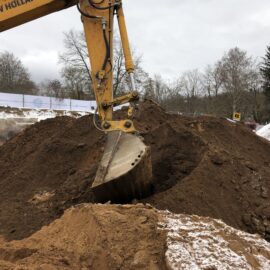 Das Bild zeigt einen großen gelben Bagger, der mit seiner Schaufel Material aus einem hohen Sandhügel entnimmt. Der Hügel besteht aus lockerem, braunem Erdmaterial, das teilweise mit Schnee bedeckt ist, was auf winterliche Bedingungen hinweist. Im Hintergrund sind kahle Bäume und ein Bauzaun zu sehen, der die Baustelle begrenzt.
