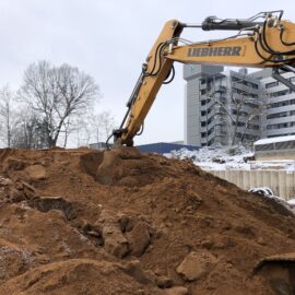 Das Bild zeigt einen großen gelben Bagger, der auf einem hohen Sandhügel arbeitet und mit seiner Schaufel Material bewegt. Der Hügel besteht aus lockerem, rötlich-braunem Sand und größeren Erdklumpen, die teilweise mit Schnee bedeckt sind. Im Hintergrund sind kahle Bäume und ein mehrstöckiges modernes Gebäude zu sehen, was auf eine Baustelle in winterlicher Umgebung hinweist.