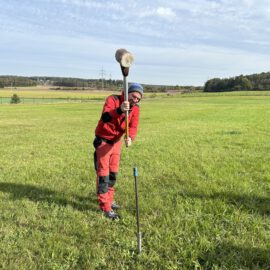 Das Bild zeigt eine Person auf einer grünen Wiese, die ein schweres Werkzeug mit beiden Händen hält und damit auf einen Metallstab oder eine Sonde schlägt. Die Person trägt rote Arbeitskleidung und steht in einer offenen Landschaft mit Feldern und Wald im Hintergrund. Der Himmel ist leicht bewölkt und vermittelt eine helle, klare Tagesstimmung.