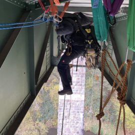 Das Bild zeigt eine Person, die unter einer großen Stahlbrücke an Seilen hängt und mit Kletterausrüstung gesichert ist. Mehrere bunte Bandschlingen und Karabiner sind an den Metallträgern der Brücke befestigt, um die Sicherung zu gewährleisten. Im Hintergrund ist ein steinerner Brückenpfeiler sowie ein bewaldetes Gebiet zu erkennen, was auf eine Übung oder Inspektion in großer Höhe hinweist.