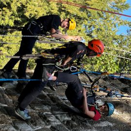 Das Bild zeigt drei Personen, die mit Kletterausrüstung und Sicherheitsseilen an einer steinernen Wand hängen. Alle tragen Helme und sind mit Karabinern, Gurten und Seilen gesichert, während sie eine Übung zur Rettung oder Sicherung durchführen. Im Hintergrund sind grüne Bäume zu sehen, und die Szene findet bei Tageslicht im Freien statt.