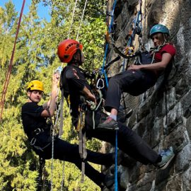 Das Bild zeigt drei Personen, die mit Kletterausrüstung und Sicherheitsseilen an einer steinernen Wand hängen. Alle tragen Helme und sind mit Karabinern, Gurten und Seilen gesichert, während sie eine Übung zur Rettung oder Sicherung durchführen. Im Hintergrund sind grüne Bäume zu sehen, und die Szene findet bei Tageslicht im Freien statt.
