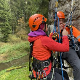Das Bild zeigt zwei Personen, die mit Kletterausrüstung und Sicherheitsseilen an einer steinernen Wand arbeiten. Beide tragen Helme und Gurte, während eine Person die Seile oder Karabiner der anderen überprüft oder justiert. Im Hintergrund ist ein bewaldetes Gebiet mit einem kleinen Bach zu sehen, was auf eine Übung im Freien hinweist.