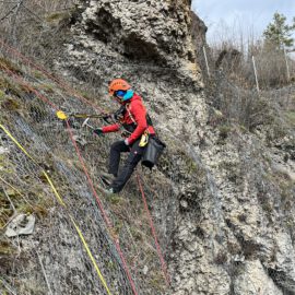 Eine Person arbeitet an einer steilen Felswand und ist mit Kletterausrüstung sowie Sicherheitsseilen gesichert. Die Felswand ist teilweise mit Drahtnetzen bedeckt, die vermutlich zur Sicherung gegen Steinschlag dienen. Im Hintergrund sind einige Bäume und ein bewölkter Himmel zu sehen.