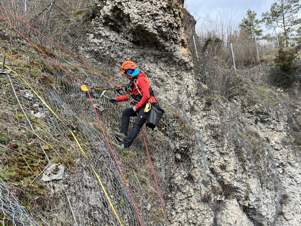Eine Person arbeitet an einer steilen Felswand und ist mit Kletterausrüstung sowie Sicherheitsseilen gesichert. Die Felswand ist teilweise mit Drahtnetzen bedeckt, die vermutlich zur Sicherung gegen Steinschlag dienen. Im Hintergrund sind einige Bäume und ein bewölkter Himmel zu sehen.