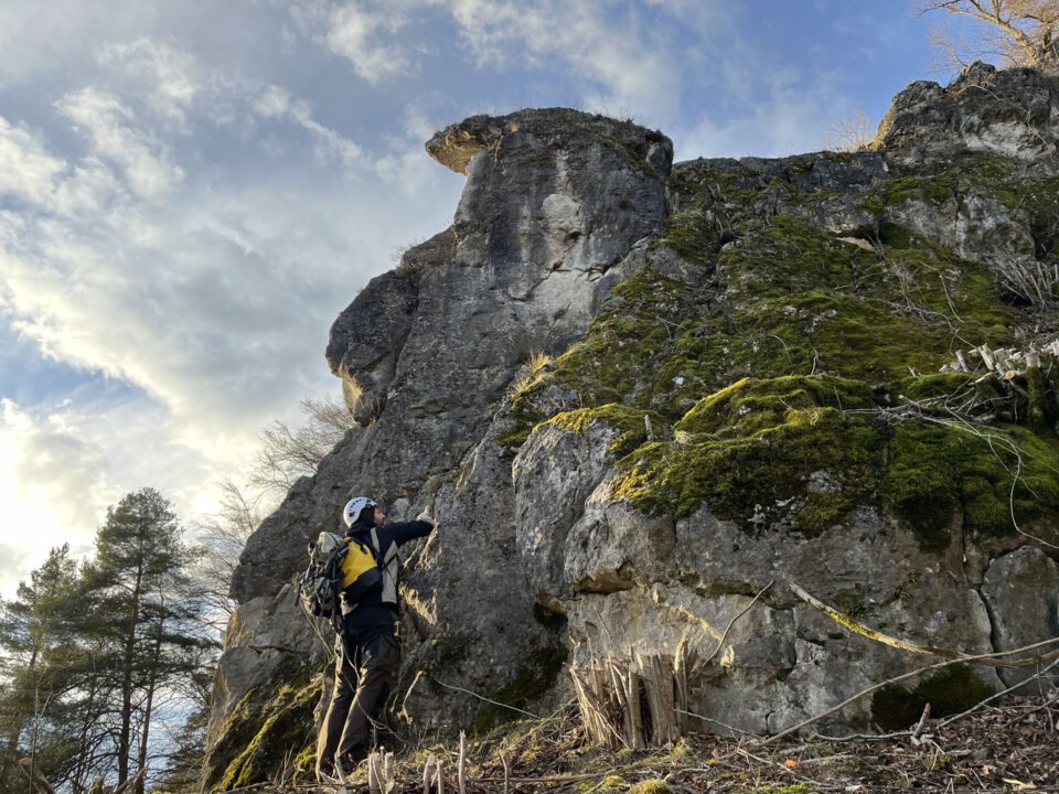 Das Bild zeigt eine steile Felswand mit markanten Strukturen und teilweise mit Moos bewachsenen Flächen. Im Vordergrund steht eine Person mit Helm und Rucksack, die nach oben blickt und die Felsformation begutachtet. Der Himmel ist teilweise bewölkt, mit Sonnenlicht, das die Felsen und die umliegende Vegetation beleuchtet, wodurch die Details der Oberfläche gut sichtbar sind.