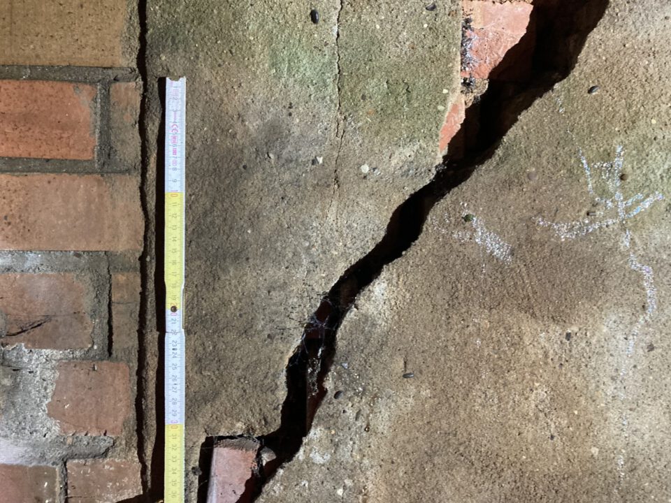 Das Bild zeigt eine Mauer aus Naturstein und Ziegeln, die einen großen, diagonalen Riss aufweist. Der Riss verläuft durch den Putz und legt an mehreren Stellen die darunterliegenden roten Ziegel frei. Links neben dem Schaden steht eine Messlatte zur Größenbestimmung, und die Oberfläche der Wand ist unregelmäßig mit Verfärbungen und leichten Abnutzungsspuren.