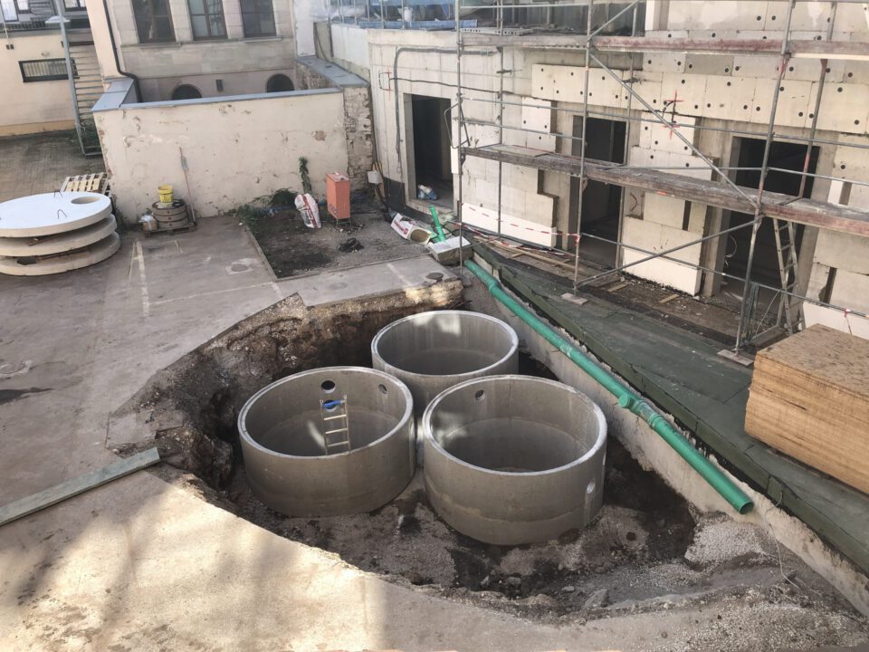 Das Bild zeigt eine Baustelle mit drei großen, runden Betonschächten, die in eine vorbereitete Grube eingebaut wurden. Die Schächte sind offen und teilweise mit Leitern ausgestattet, was auf Arbeiten im Inneren hinweist. Im Hintergrund befindet sich ein Gebäude mit Gerüst an der Fassade, sowie Baumaterialien und Rohre, die auf eine Sanierungs- oder Installationsmaßnahme hindeuten.