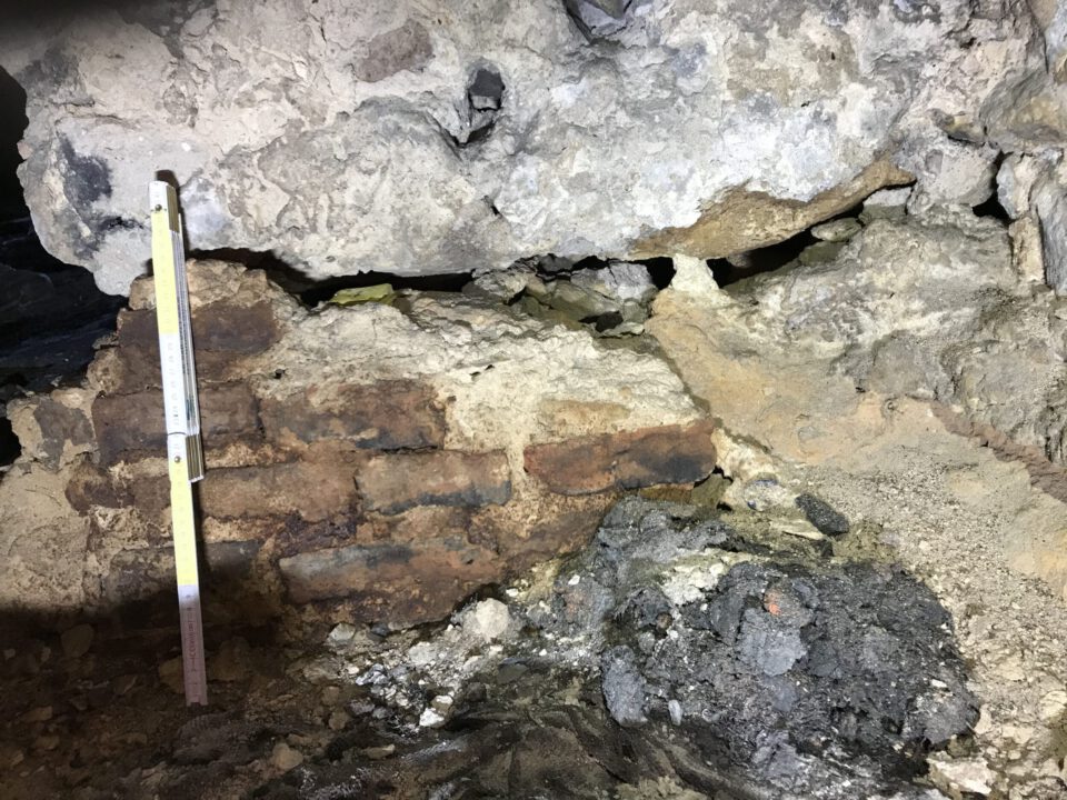 Das Bild zeigt eine beschädigte Mauer aus Naturstein und Ziegeln, die deutliche Abnutzungsspuren aufweist. Die Oberfläche ist unregelmäßig, mit Rissen und Ausbrüchen, und im unteren Bereich liegen lose Steine sowie dunkles, bröckeliges Material. Links steht eine Messlatte zur Größenbestimmung, die an die Mauer angelehnt ist, während die Umgebung von Erd- und Staubablagerungen geprägt ist.