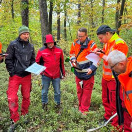 Das Bild zeigt eine Gruppe von sechs Personen, die in einem Wald zusammenstehen. Die Personen tragen überwiegend orangefarbene Warnjacken, einige auch rote oder dunkle Kleidung, und halten Unterlagen sowie Tablets in den Händen. Der Boden ist mit grüner Vegetation bedeckt, und im Hintergrund sind zahlreiche Bäume mit herbstlich gefärbten Blättern zu sehen.