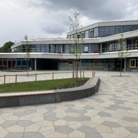 Das Bild zeigt einen modernen Schulhof mit einem großen, gepflasterten Platz, der aus sechseckigen Steinplatten besteht. In der Mitte befindet sich eine Grünfläche mit jungen Bäumen, die von einem niedrigen Holzgeländer umgeben ist. Entlang der Grünfläche sind geschwungene Sitzbänke integriert. Im Hintergrund steht ein mehrstöckiges Schulgebäude mit großen Glasfronten und Balkonen, dessen Fassade in hellen und grauen Farbtönen gestaltet ist. Mehrere Fensterrahmen sind farbig abgesetzt, was dem Gebäude einen lebendigen Charakter verleiht.