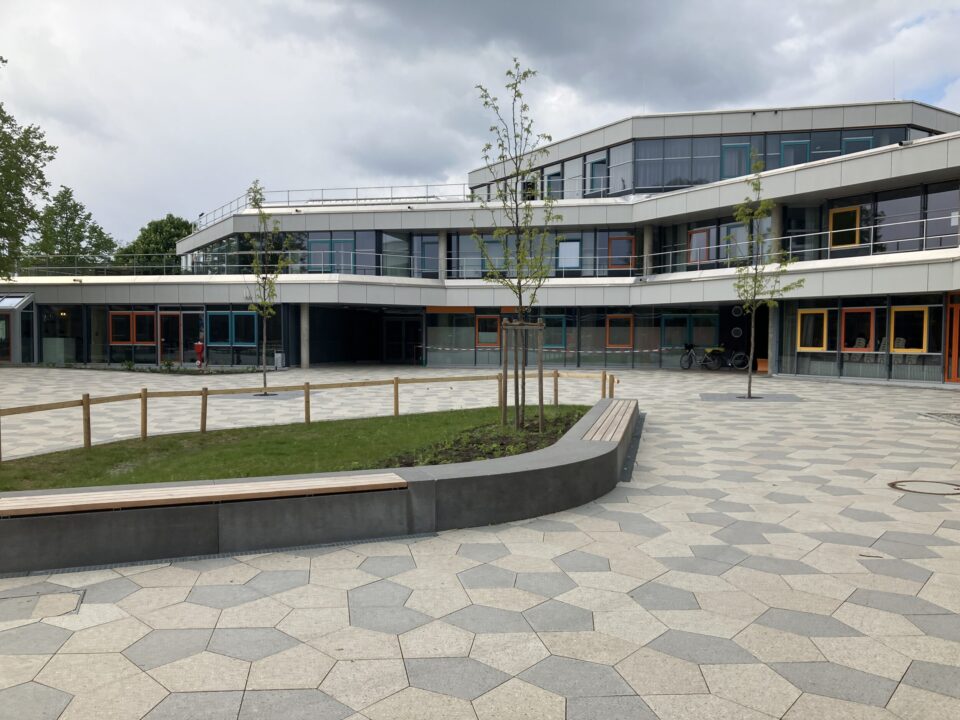 Das Bild zeigt einen modernen Schulhof mit einem großen, gepflasterten Platz, der aus sechseckigen Steinplatten besteht. In der Mitte befindet sich eine Grünfläche mit jungen Bäumen, die von einem niedrigen Holzgeländer umgeben ist. Entlang der Grünfläche sind geschwungene Sitzbänke integriert. Im Hintergrund steht ein mehrstöckiges Schulgebäude mit großen Glasfronten und Balkonen, dessen Fassade in hellen und grauen Farbtönen gestaltet ist. Mehrere Fensterrahmen sind farbig abgesetzt, was dem Gebäude einen lebendigen Charakter verleiht.