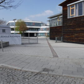 Das Bild zeigt den Eingangsbereich der Realschule Puchheim. Im Vordergrund befindet sich eine gepflasterte Fläche mit einem breiten Gehweg und einem Steinblock, auf dem der Schriftzug Realschule Puchheim zu lesen ist. Rechts steht ein Gebäude mit einer Holzfassade und großen Fenstern, während im Hintergrund ein moderner Schulkomplex mit Glasfronten und mehreren Etagen sichtbar ist. Links sind ein Baum und ein kleiner Grünbereich zu erkennen, der den Platz auflockert.