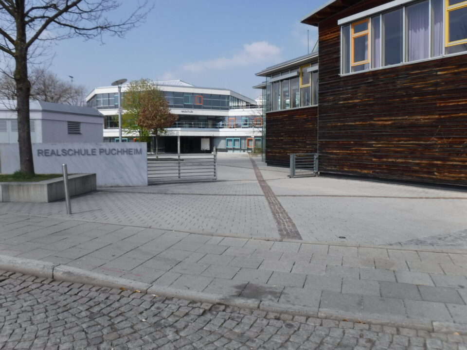 Das Bild zeigt den Eingangsbereich der Realschule Puchheim. Im Vordergrund befindet sich eine gepflasterte Fläche mit einem breiten Gehweg und einem Steinblock, auf dem der Schriftzug Realschule Puchheim zu lesen ist. Rechts steht ein Gebäude mit einer Holzfassade und großen Fenstern, während im Hintergrund ein moderner Schulkomplex mit Glasfronten und mehreren Etagen sichtbar ist. Links sind ein Baum und ein kleiner Grünbereich zu erkennen, der den Platz auflockert.
