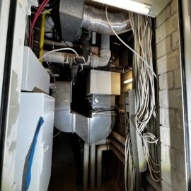 Das Bild zeigt einen engen Technikschacht mit zahlreichen Installationen und Leitungen. Im Vordergrund sind viele weiße Kabelbündel zu sehen, die an der rechten Wand entlanggeführt werden. Im hinteren Bereich befinden sich metallische Lüftungskanäle, isolierte Rohre und verschiedene technische Komponenten, darunter ein rechteckiges Gerät mit silberner Oberfläche. An der Decke ist eine Leuchtstofflampe angebracht, die den Schacht ausleuchtet. Die Wände bestehen aus Mauerwerk, und insgesamt wirkt die Umgebung wie ein Bereich für Gebäudetechnik oder Haustechnik.