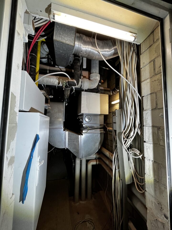 Das Bild zeigt einen engen Technikschacht mit zahlreichen Installationen und Leitungen. Im Vordergrund sind viele weiße Kabelbündel zu sehen, die an der rechten Wand entlanggeführt werden. Im hinteren Bereich befinden sich metallische Lüftungskanäle, isolierte Rohre und verschiedene technische Komponenten, darunter ein rechteckiges Gerät mit silberner Oberfläche. An der Decke ist eine Leuchtstofflampe angebracht, die den Schacht ausleuchtet. Die Wände bestehen aus Mauerwerk, und insgesamt wirkt die Umgebung wie ein Bereich für Gebäudetechnik oder Haustechnik.