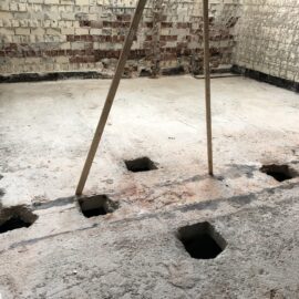 Das Bild zeigt einen Raum mit freigelegtem Betonboden, in dem mehrere quadratische Öffnungen in den Boden eingelassen sind. Zwei Holzstützen stehen mittig im Raum und stützen offenbar die Decke. Die Wände bestehen aus altem Mauerwerk mit teilweise abgeplatztem Putz, was auf Renovierungs- oder Umbauarbeiten hinweist.