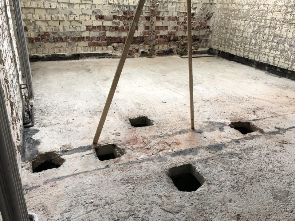 Das Bild zeigt einen Raum mit freigelegtem Betonboden, in dem mehrere quadratische Öffnungen in den Boden eingelassen sind. Zwei Holzstützen stehen mittig im Raum und stützen offenbar die Decke. Die Wände bestehen aus altem Mauerwerk mit teilweise abgeplatztem Putz, was auf Renovierungs- oder Umbauarbeiten hinweist.