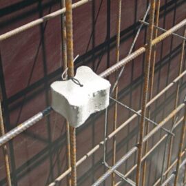 Das Bild zeigt eine Bewehrungskonstruktion aus mehreren gerippten Stahlstäben, die ein Gitter bilden. In der Mitte ist ein Abstandshalter aus hellem Material befestigt, der eine kreuzförmige Form hat und mit einem Draht an den Stäben fixiert ist.