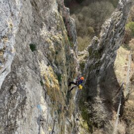 Das Bild zeigt eine steile Felswand, an der eine Person mit Kletterausrüstung gesichert hängt. Die Person trägt einen Helm und hat ein gelbes Seil oder einen Sack bei sich, während oberhalb ein Sicherungsseil verläuft. Im Hintergrund sind weitere schmale Felsformationen sowie bewaldetes Gelände zu sehen, was auf eine natürliche, bergige Umgebung hinweist.