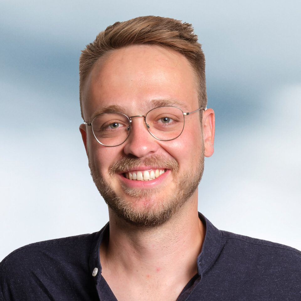 Das Bild zeigt ein professionelles Porträt eines jungen Mannes mit kurzem, nach oben gestyltem hellbraunem Haar, runder Brille und leichtem Bart. Er lächelt offen in die Kamera und trägt ein dunkles, schlichtes Hemd. Der Hintergrund ist weich verlaufend in hellen Blau- und Weißtönen gehalten und wirkt ruhig und neutral.