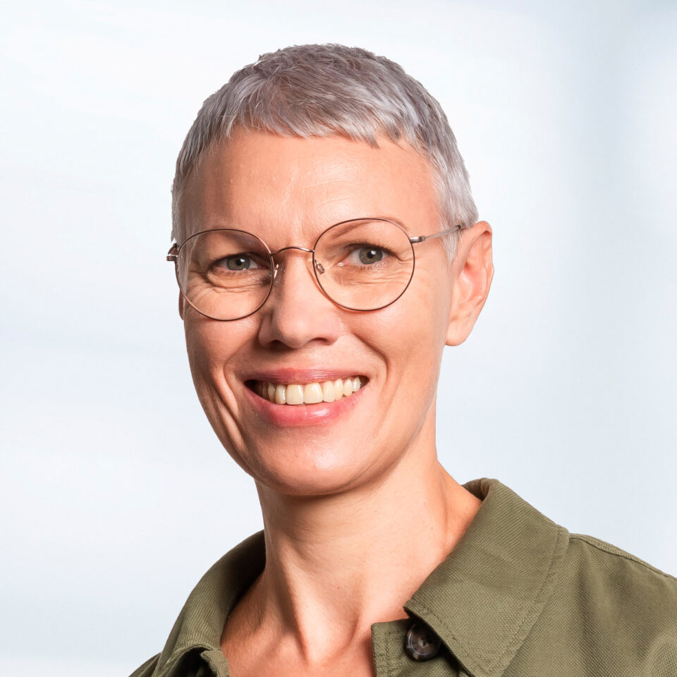 Das Bild zeigt ein professionelles Porträt einer Frau mit sehr kurzem, grauem Haar und einer runden Brille, die freundlich in die Kamera lächelt. Sie trägt ein olivgrünes Oberteil mit Kragen, das einen schlichten und gepflegten Eindruck macht. Der Hintergrund ist weich verlaufend in hellen Grau- und Blautönen gehalten und sorgt für eine ruhige, klare Bildwirkung.