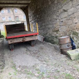 Das Bild zeigt einen überdachten Durchgang mit groben Steinwänden und einem Holzdach. In der Mitte steht ein alter Anhänger mit rotem Rahmen und Holzaufbau, der auf zwei Rädern ruht. Rechts neben dem Anhänger befindet sich ein rostiges Metallfass auf dem Boden, und durch den Torbogen im Hintergrund ist ein Gebäude mit mehreren Fenstern sichtbar.