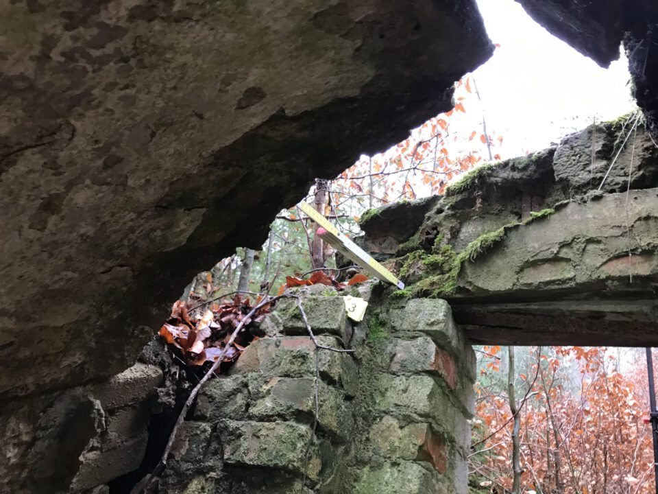 Das Bild zeigt ein stark beschädigtes steinernes Eingangsportal in einer bewaldeten Umgebung. Von unten blickt man auf eine aufgebrochene, schräg liegende massive Deckenplatte, die über dem Eingang stark gerissen und teilweise abgesackt ist. Die Mauerpfeiler bestehen aus unregelmäßigem Bruchsteinmauerwerk, das deutlich verwittert ist. Zwischen den Steinen sind Moos, Erdreich und Laub sichtbar. Auf dem oberen Mauerbereich liegt ein gelber Zollstock, daneben ist eine kleine gelbe Markierung mit der Zahl „3“ befestigt. Lose Äste und Herbstlaub haben sich auf der beschädigten Struktur gesammelt.
