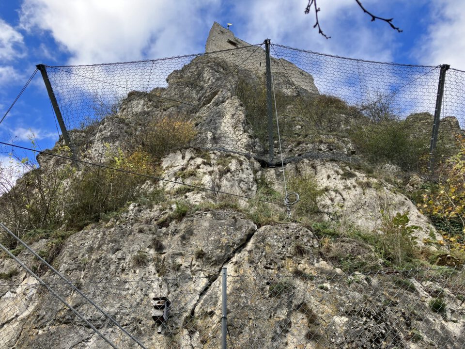 Das Bild zeigt eine steile Felswand, die mit einem Steinschlagschutznetz gesichert ist. Das Netz ist an mehreren stabilen Metallpfosten befestigt und wird zusätzlich durch Stahlseile verstärkt, die quer über die Felsfläche verlaufen. Zwischen den Felsen wachsen kleine Sträucher und Pflanzen, während im Hintergrund ein blauer Himmel mit einigen weißen Wolken zu sehen ist.