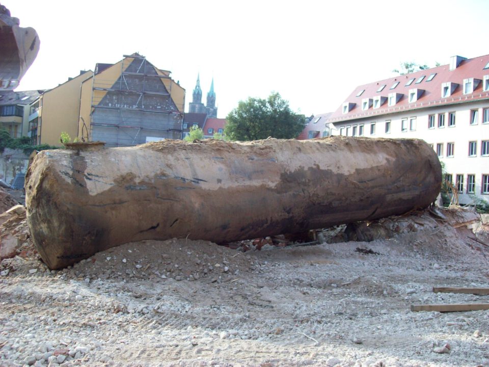 Das Bild zeigt einen großen, zylindrischen Tank, der aus dem Erdreich ausgebaut wurde und nun auf einer Baustelle liegt. Der Tank ist stark verschmutzt und mit Erd- und Rostspuren bedeckt, was auf eine lange Lagerung im Boden hinweist. Im Hintergrund sind mehrstöckige Gebäude und ein teilweise abgerissenes Haus zu sehen, sowie die Türme einer Kirche, die über die Dächer hinausragen.