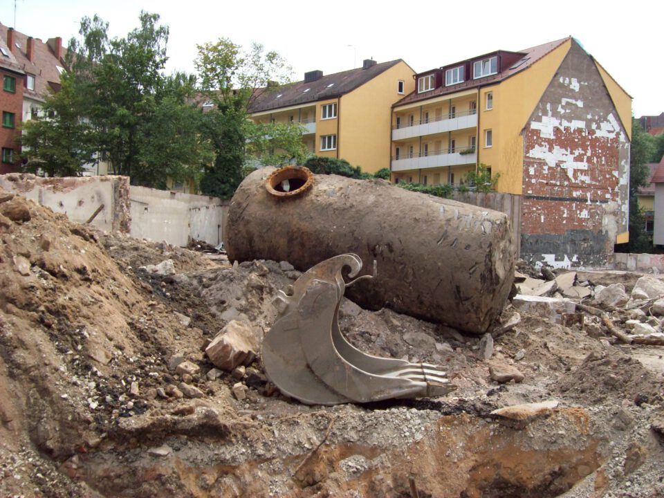 Das Bild zeigt eine Baustelle mit einem großen, zylindrischen Tank, der aus dem Erdreich ausgebaut wurde und schräg auf einem Erdhaufen liegt. Der Tank ist stark verschmutzt und weist Rost- und Erdspuren auf, zudem ist eine runde Öffnung an der Oberseite sichtbar. Im Vordergrund liegen mehrere große Metallteile, vermutlich Baggerschaufeln, und im Hintergrund sind mehrstöckige Wohnhäuser sowie eine teilweise beschädigte Fassade zu sehen.