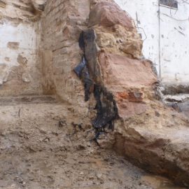 Das Bild zeigt eine freigelegte Mauer aus grob behauenen Sandsteinblöcken und Ziegeln, die teilweise beschädigt ist. An der Wand ist eine dunkle, teerartige Abdichtung zu erkennen, die in Streifen von oben nach unten verläuft. Der Boden davor besteht aus feuchtem Erdreich und wirkt uneben, während die Hintergrundwand stark verwittert und fleckig ist.